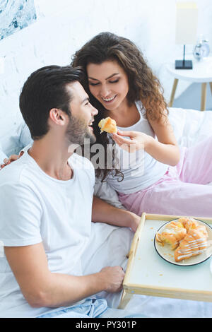 Happy young woman smiling alimentation petit ami avec un croissant au lit Banque D'Images