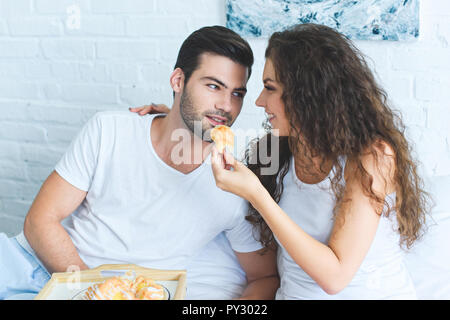 Belle jeune femme souriante d'alimentation petit ami avec un croissant au lit Banque D'Images