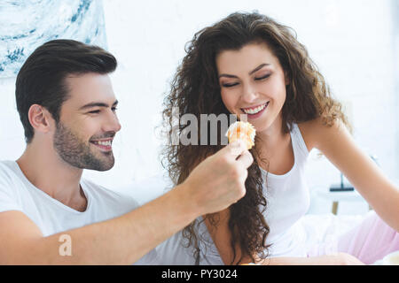 Jeune homme souriant heureux alimentation petite amie avec des croissants pour le petit-déjeuner dans la chambre Banque D'Images