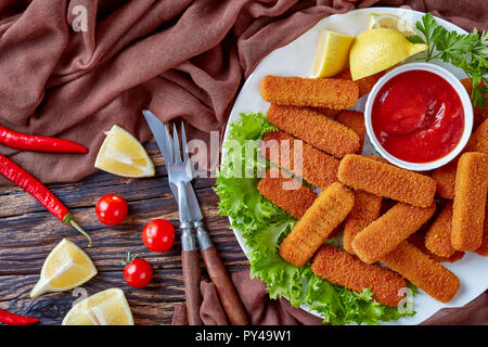 Vue supérieure de l'crumbed les bâtonnets de poisson servi sur une plaque blanche avec du citron, des feuilles de laitue et de la sauce tomate sur une vieille table rustique, vue horizontale à partir de Banque D'Images