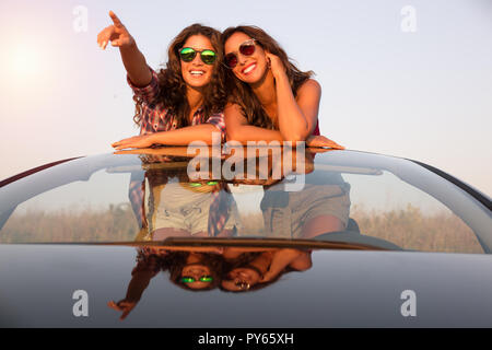 Deux belles jeunes filles avec des lunettes de soleil dans une voiture décapotable regarde quelque chose au coucher du soleil Banque D'Images