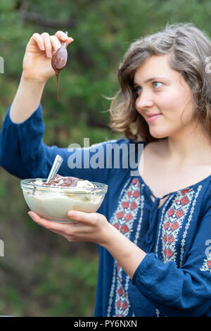 Jeune femme à l'extérieur, à l'extérieur, tenant bol en verre composé de végétalien premières, glace vanille, sauce chocolat ajout de liquide à la cuillère, looking up Banque D'Images