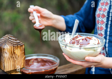 Libre de jeune femme à l'extérieur, à l'extérieur, tenant bol en verre composé de végétalien premières, glace vanille, sauce au chocolat ajouter la cuillère de liquide Banque D'Images