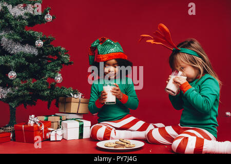 Deux petites filles, assis près d'un arbre de Noël lait de consommation. Deux enfants de manger des cookies avec du lait, assis près d'un arbre de Noël décoré. Banque D'Images