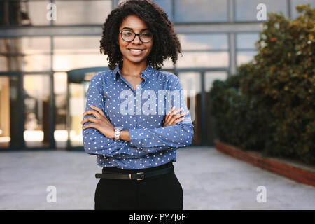 Portrait d'une jeune femme africaine positive debout dehors, les bras croisés. professionnel des affaires debout en toute confiance avec l'immeuble de bureaux dans ba Banque D'Images