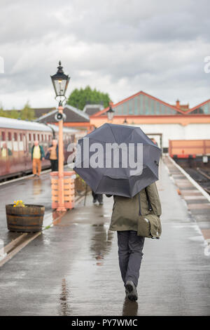 Vue arrière d'une rames féminine en anorak, tenant un parapluie à l'écart de la caméra le long de la plate-forme de chemin de fer à la gare d'époque, au Royaume-Uni, sous la pluie. Banque D'Images