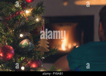 Vue de derrière du jeune homme assis devant le foyer avec le feu et l'arbre de Noël à côté de lui. Banque D'Images