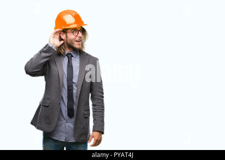 Beau jeune homme architecturaux aux cheveux longs portant un casque de sécurité sur fond isolé smiling with hand sur l'écoute d'une oreille à l'audience ou de rumeur Banque D'Images