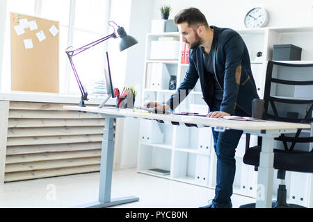 Le jeune homme se tient dans le bureau près de la table et se penche sur le moniteur. Banque D'Images