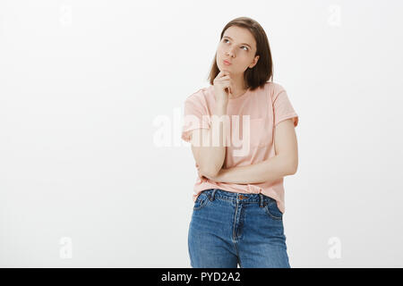 Jeune fille se demande si elle peut se permettre une plus de t-shirt. Déterminé et créatif cute woman, debout dans une pose, pensant alors que les lèvres de pliage et à la recherche dans le coin supérieur gauche, curieusement touchant chin Banque D'Images