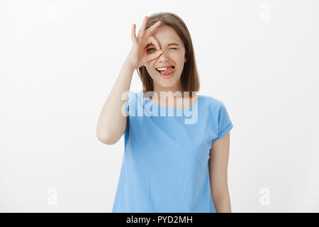 À la fille sur le côté lumineux. Joyeux positif étudiante en tee-shirt bleu, montrant bien ou sur ok et l'œil sticking out tongue, debout et heureux, heureux d'avoir du bon temps avec des amis Banque D'Images