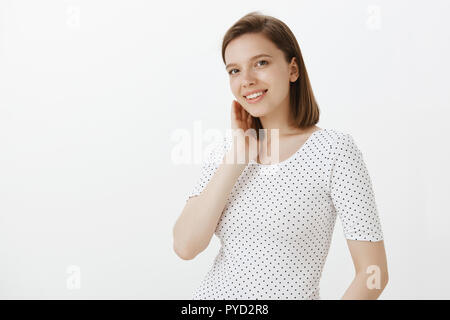 Mignon et heureux d'un collègue femelle en T-shirt, touchant doucement le cou et sourit joyeusement, tandis que l'article retourné demi-mur gris, sa timidité et le rougissement de compliment Banque D'Images
