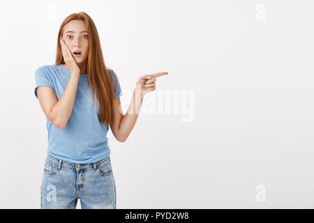 Portrait de choqué et surpris young redhead lady avec rousseur halètement bouche ouverture toucher joue et orientée vers la droite en question et curieux tandis que témoins de scène choquante Banque D'Images