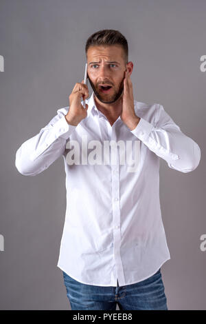 Surpris un beau jeune homme dans une chemise blanche à parler sur son téléphone et, debout devant un fond gris dans le studio. Banque D'Images