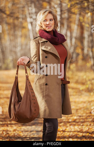 Happy young blonde woman fashion avec sac à main élégant en plein air randonnée modèle féminin portant manteau beige classique et bourgogne écharpe Banque D'Images