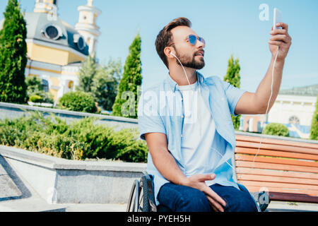 Bel homme en fauteuil roulant en tenant avec selfies smartphone et écouter de la musique sur la rue Banque D'Images