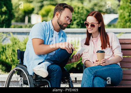 Bel Ami en fauteuil roulant montrant quelque chose sur smartphone pour petite amie sur banc Banque D'Images