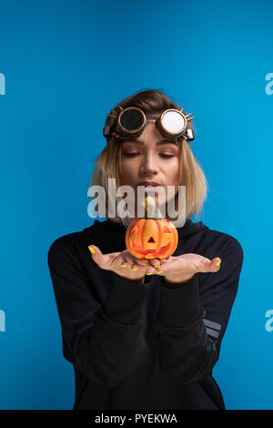 Femme portant des lunettes steampunk et black hoodie tenant une Halloween citrouille sculptée. Steampunk goth à thème tournage en studio contre fond bleu Banque D'Images