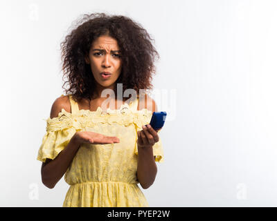 Jolie jeune femme afro-américaine en jaune dress holding petite boîte de bijoux sur fond blanc. Déçu upset girl, elle n'aime pas cadeau. Banque D'Images