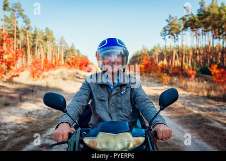 Senior man riding scooter sur la route forestière de l'automne. Heureux conducteur en équitation casque scooter. Banque D'Images