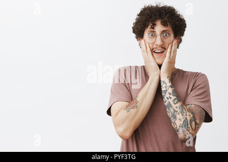 Jusqu'à la taille de shto excité et heureux amusé beau jeune homme hipster étudiant dans brown t-shirt avec des tatouages de la moustache et des cheveux bouclés sombres lunettes en tenant la main sur le visage et le sourire de surprise Banque D'Images