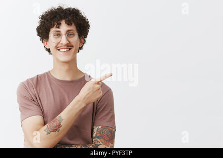Vous ne trouverez pas meilleur endroit pour la publicité. Friendly beau et jeune homme élégant modèle avec curly hairstyle tatouages et moustache souriant joyeusement en montrant le coin supérieur droit Banque D'Images