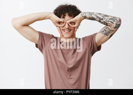 Jusqu'à la taille de balle drôle et espiègle jeune homme aux cheveux bouclés avec bras tatoués et moustache faire un masque pour les yeux avec les doigts tout sourire, de jouer et s'amuser sur fond gris Banque D'Images