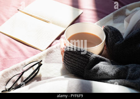 Le café et lire au lit le matin, se prélasser au soleil. Les mains tenant tasse fumante de chocolat chaud lire au lit Banque D'Images