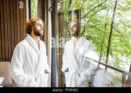 Bel homme en peignoir, debout près de la fenêtre avec vue magnifique sur la forêt dans la chambre d'hôtel moderne
