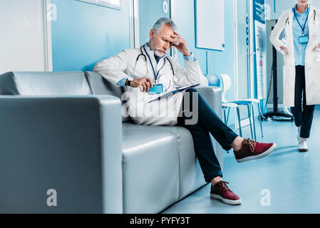 Médecin homme d'âge moyen réfléchi avec presse-papiers assis sur un canapé, tandis que son collègue marchant derrière in hospital corridor Banque D'Images