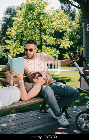 Rire femme couchée sur mari jambes près de landau dans park et reading book Banque D'Images