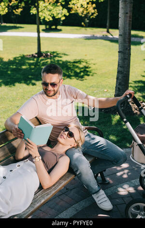 Portrait de femme couchée sur mari jambes près de landau dans park et holding book Banque D'Images