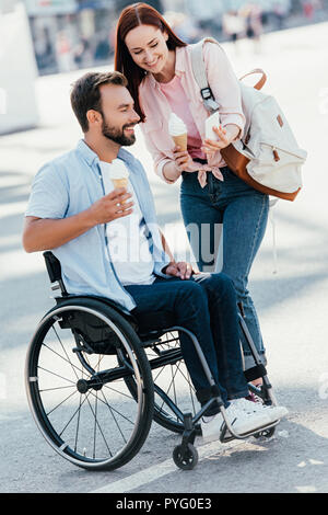 Amie avec la crème glacée montrant quelque chose sur smartphone pour bel ami en fauteuil roulant sur la rue Banque D'Images