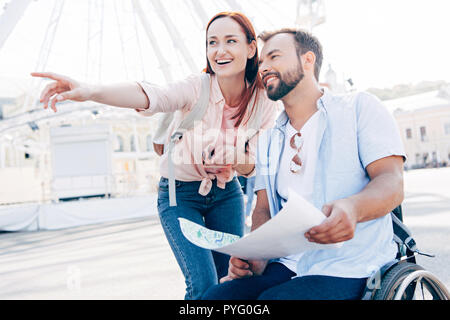 Smiling handsome boyfriend in wheelchair holding la carte et petite amie pointant sur quelque chose sur la rue Banque D'Images