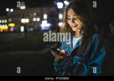 Belle jeune fille texting on cell phone piscine plus floue nuit rue background, selective focus Banque D'Images