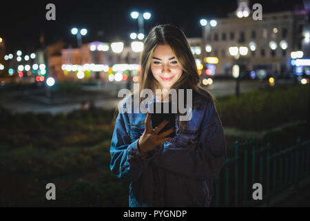 Belle jeune fille texting on cell phone piscine plus floue nuit rue background, selective focus Banque D'Images