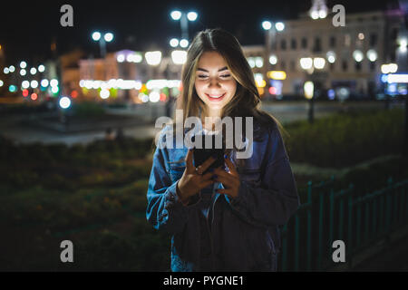 Belle jeune fille texting on cell phone piscine plus floue nuit rue background, selective focus Banque D'Images