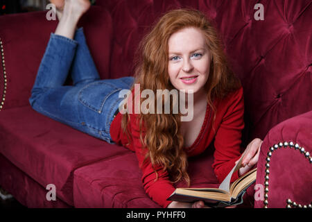 Riche jeune rousse femme lisant un livre sur un canapé de velours rouge Banque D'Images