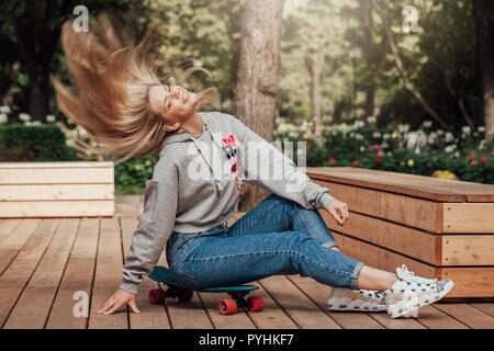 Jeune femme en jeans assis sur la planche à roulettes en parc. Style de vie. Les poils de vol Banque D'Images