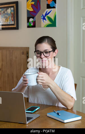 Jeune femme artiste regardant le coffre tenant une chope de prendre un verre avec les périphériques de communication de son travail table de salle à manger à la maison. Banque D'Images