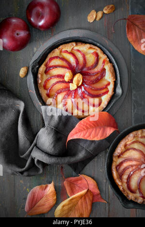 Crumble aux prunes tartes faites maison cuit dans des tranches de petits poêlons en fer. Haut de jeter sur conseil en bois brun foncé avec un peu de pruneau, noix et feuilles rouges Banque D'Images