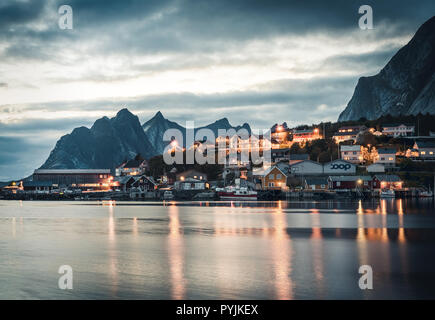 Village de la pêche norvégienne Reine à l'îles Lofoten en Norvège. Nuages coucher de soleil spectaculaire en mouvement sur des sommets de montagnes, dans le crépuscule. Reflet de stree Banque D'Images
