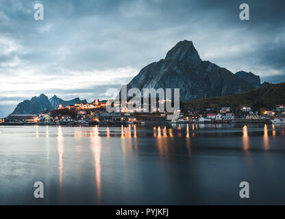 Village de la pêche norvégienne Reine à l'îles Lofoten en Norvège. Nuages coucher de soleil spectaculaire en mouvement sur des sommets de montagnes, dans le crépuscule. Reflet de stree Banque D'Images