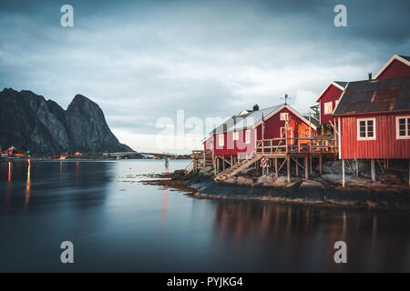 Village de la pêche norvégienne Reine à l'îles Lofoten en Norvège. Nuages coucher de soleil spectaculaire en mouvement sur des sommets de montagnes, dans le crépuscule. Reflet de stree Banque D'Images