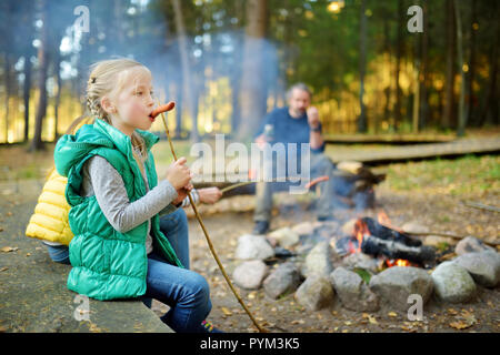 Jolie petite soeurs et leur père le rôtissage des hot-dogs sur des bâtons à feu. Les enfants s'amuser au feu de camp. Camping avec les enfants dans la forêt de l'automne. Famille Banque D'Images