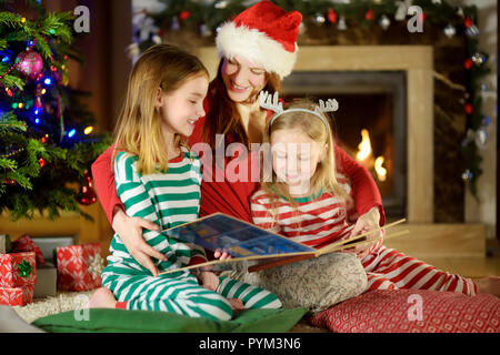 Jeune mère et ses deux petites filles de la lecture d'un livre d'un arbre de Noël dans la salle de séjour en hiver Banque D'Images