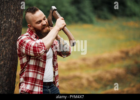 Lumberjack homme avec barbe et moustache en chemise à carreaux rouge avec lame de hache se rase sur fond naturel Banque D'Images