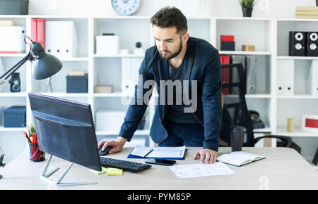 Le jeune homme se tient dans le bureau près de la table et se penche sur le moniteur. Banque D'Images
