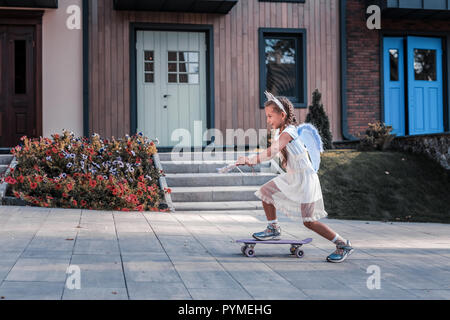 Dark-haired little girl wearing white dress et fairy wings skateboarding Banque D'Images