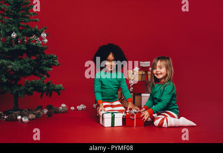 Les petites filles jouer avec leurs cadeaux de Noël assis sur le plancher. Happy kids multi ethnic avec leurs cadeaux de Noël assis à côté d'un arbre de Noël. Banque D'Images